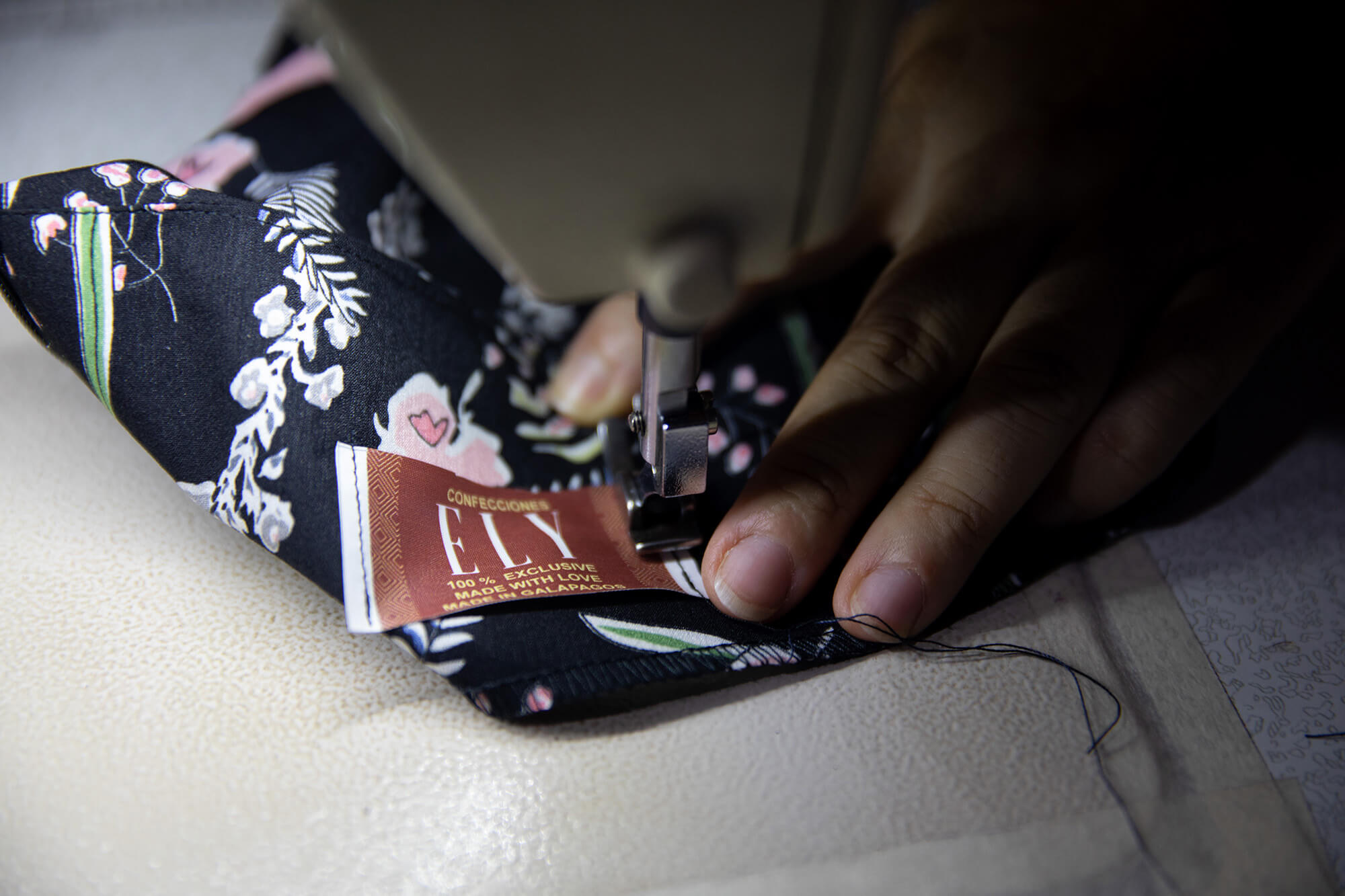 Close up of a sewing machine. Alava’s hand guides the edge of a pair of pants under the needle to stitch in her label, which reads “Confecciones Ely.”