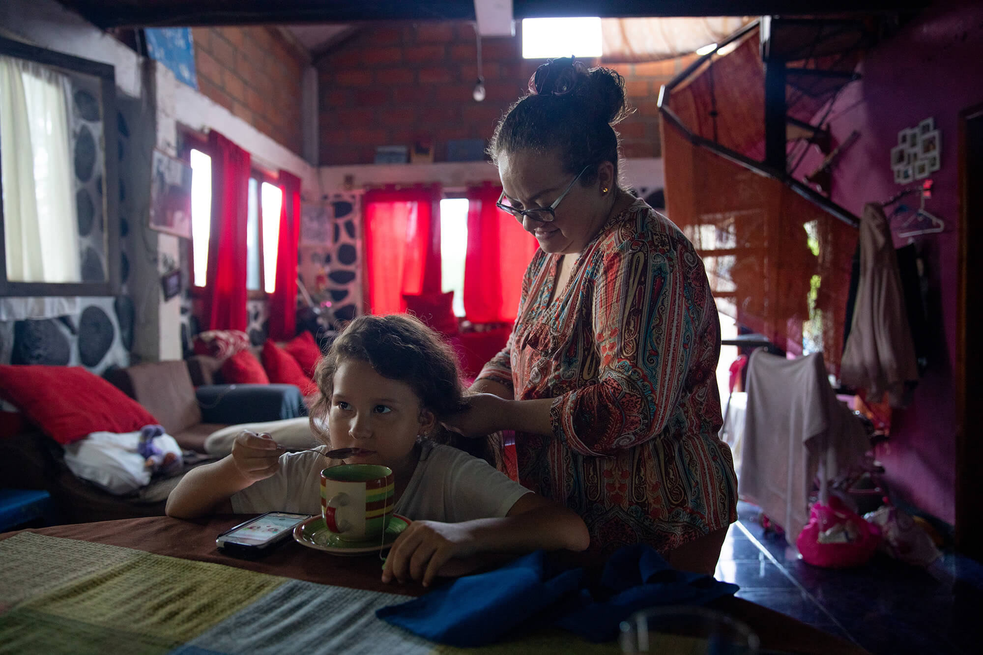 Alava, right, works her hands through daughter Amaya’s hair (left). Both are inside Alava’s home; there are red curtains in the background and a spiral staircase to Alava’s right. Amaya is sitting and sipping tea from a spoon. She wears a white shirt. Alava is wearing a red, patterned shirt and is standing.