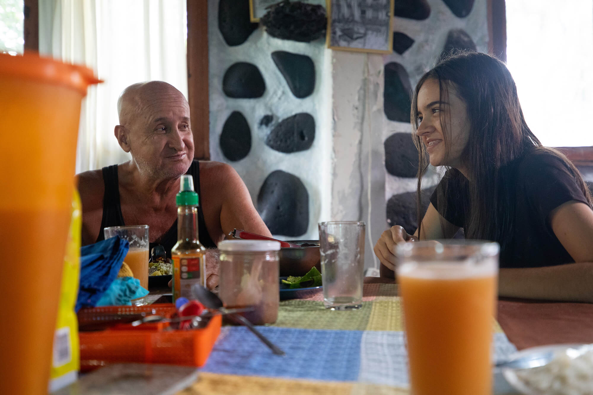 Germán, Alava’s husband (left), and her daughter, Laura, smile at each other as they sit at the family’s kitchen table for lunch. In the foreground, there is a cup of juice and an orange container. Germán is wearing a black tank top, and Laura is wearing a black shirt. Behind them, the white walls, dotted with black volcanic rock, can be seen. A drawing of a boat hangs above Germán and Laura on the wall.