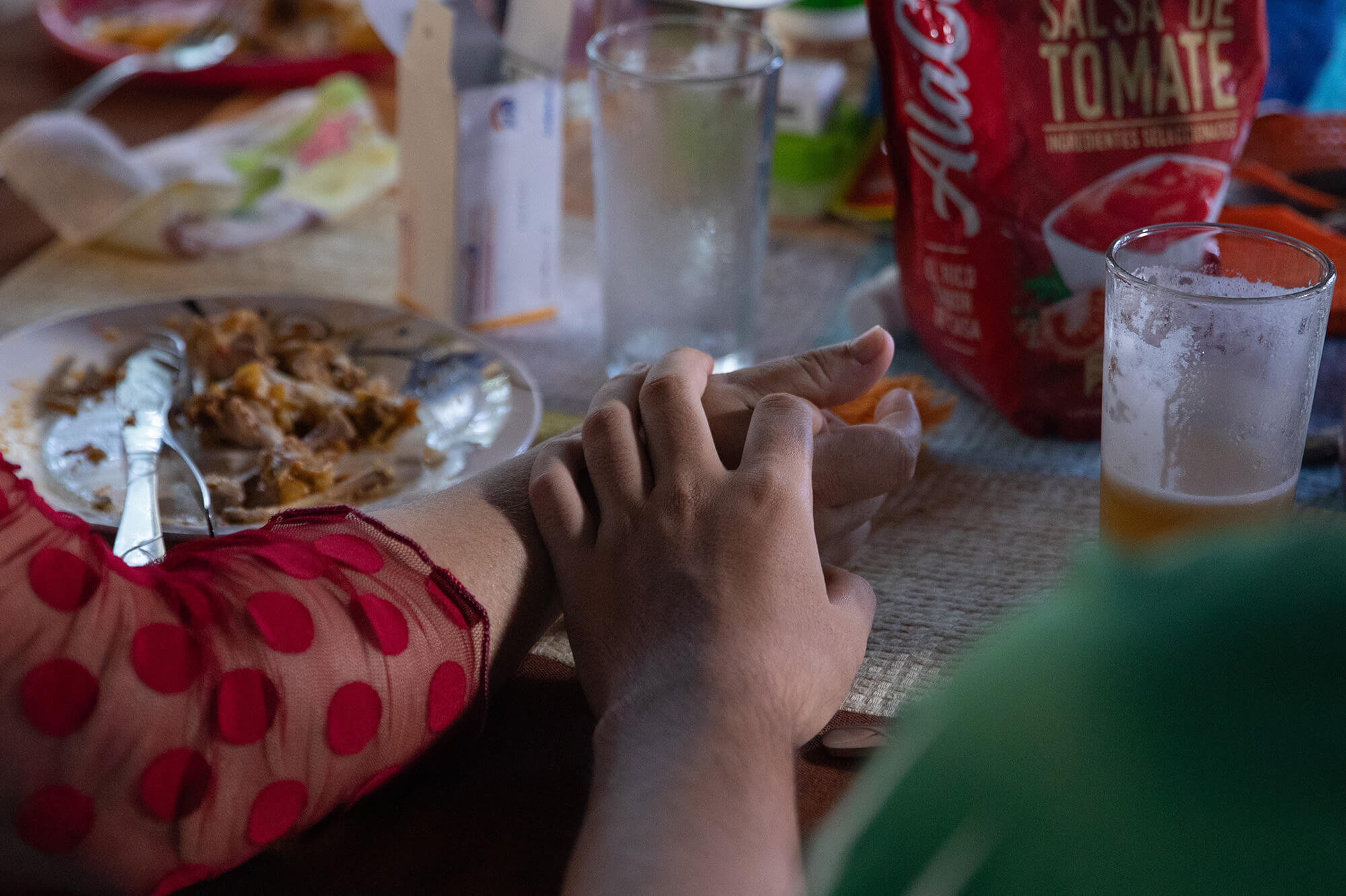 Alava, left, and her son Nicholas hold hands at the family kitchen table. A close up image, Alava’s arm has a red sleeve with red polka dots, and a plate with food is to her left. At right is a cup with juice in it and a bag of ketchup.