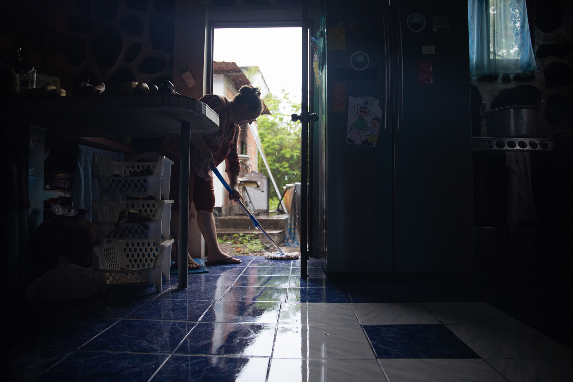 Alava mops her blue, tile kitchen floor as rain falls in the background. Alava is framed by a doorway that leads outside. To the right of the image is the kitchen fridge, which is silver, and to the left is a countertop.