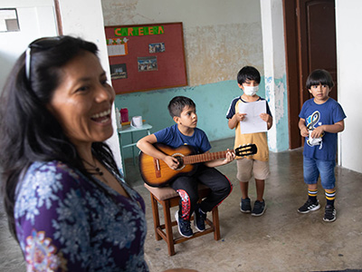Campués smiles as she watches her students perform. Three students perform next to her with one of them playing guitar. 
