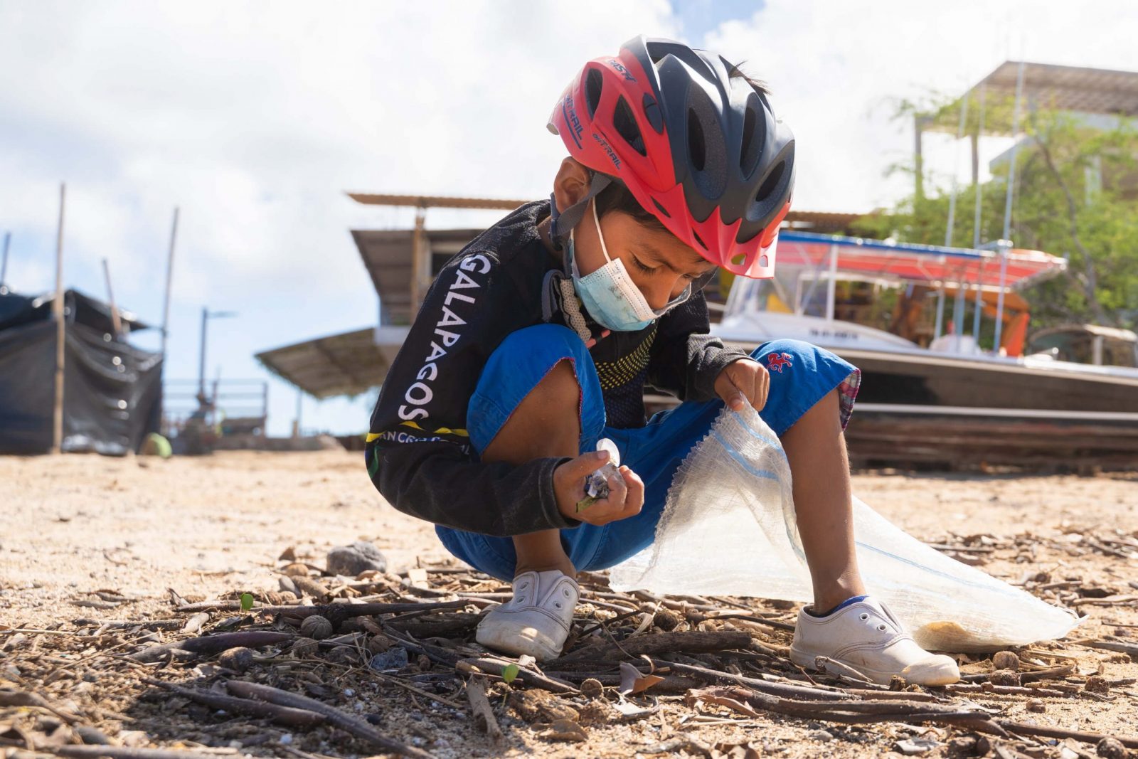 A little boy, Justin, wearing a red bike helmet squats on the beach with a handful of plastics in one hand and a reusable trash bag in the other.