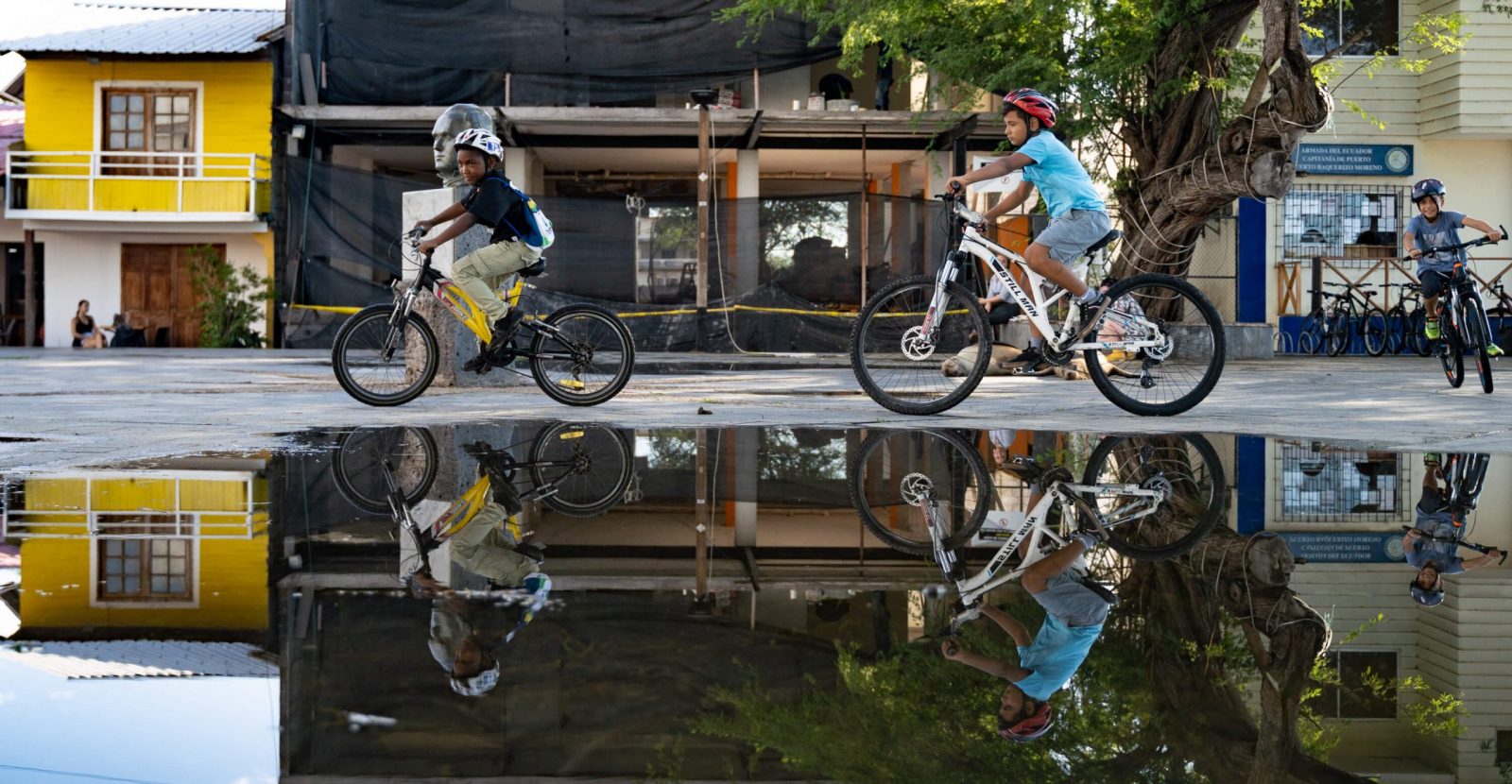 Three children ride on their bikes in the plaza right behind a large puddle that reflects their images.
