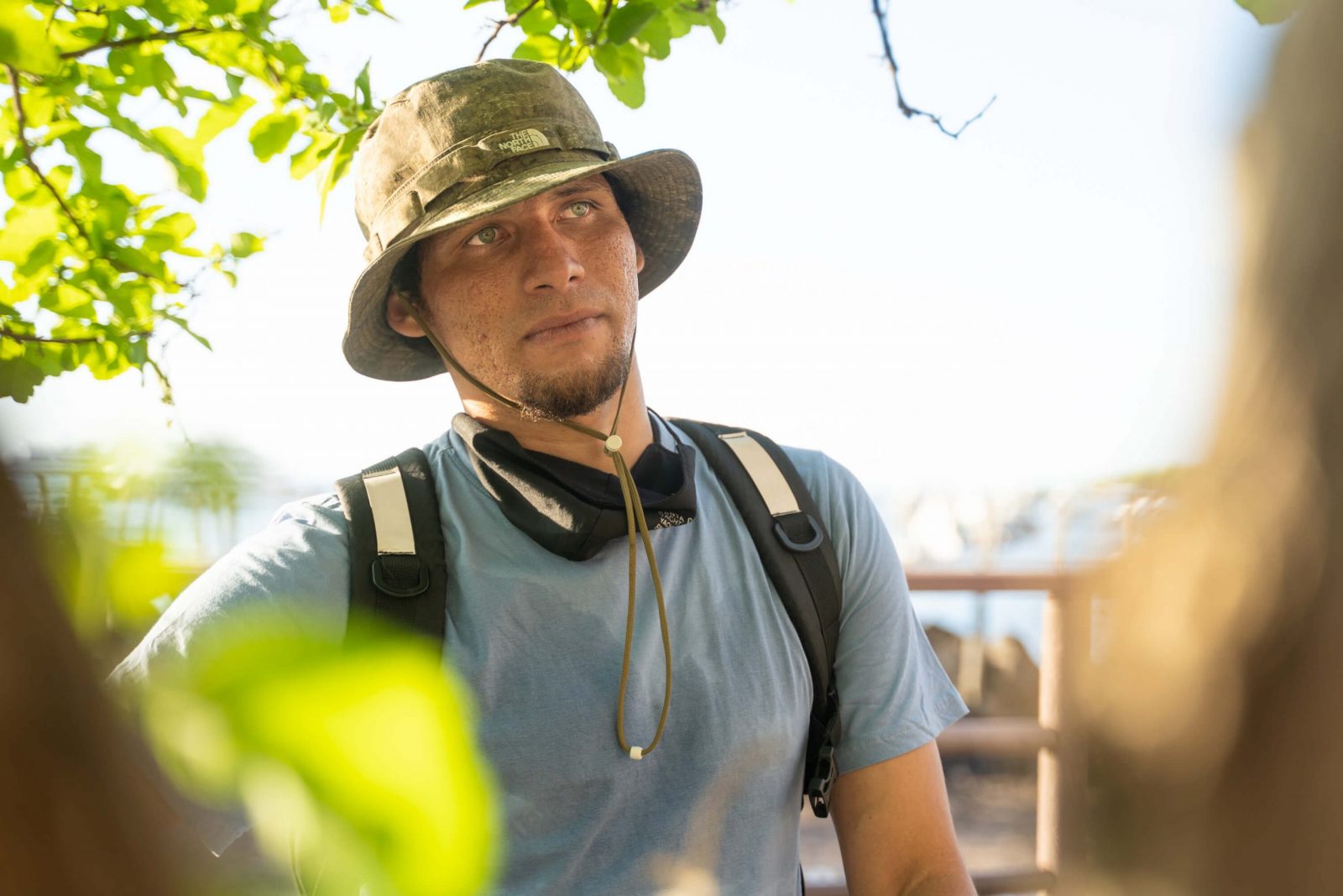 Jaison, wearing a green bucket hat, blue t-shirt, and backpack gazes at a tourist with his green eyes.