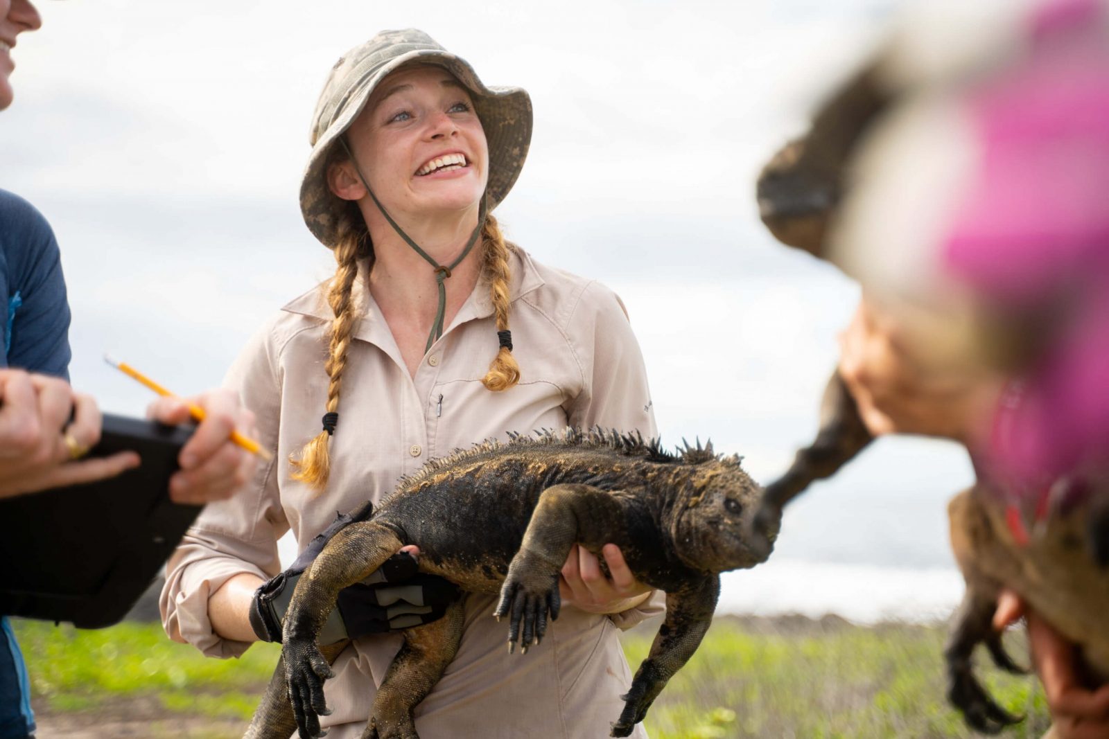 A female researcher wearing a camouflaged-patterned hat smiles at her colleagues as she holds a marine iguana.